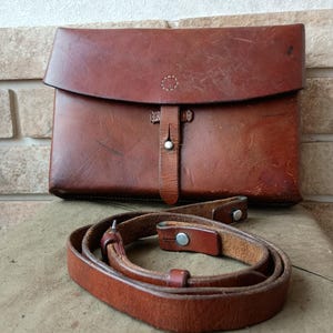 Puede incluir: Una cartera de cuero marrón con cierre de solapa y una correa para el hombro a juego. La cartera tiene forma rectangular con cierre de botón. La correa es ajustable y tiene herrajes metálicos.