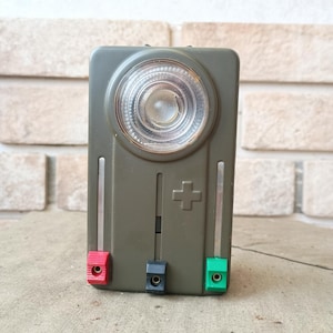 Op de afbeelding: Een olijfgroene vintage zaklamp met een cirkelvormige lens en een plusteken. De zaklamp heeft rode, zwarte en groene aansluitingen. De achtergrond is een bakstenen muur.