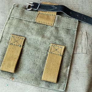Könnte beinhalten: Eine quadratische olivgrüne Segeltuchtasche mit zwei vertikalen beigefarbenen Segeltuchschlaufen auf der Vorderseite und einem schwarzen Lederriemen mit Metallschnalle an der Oberkante. Die Tasche besteht aus strapazierfähigem Stoff mit sichtbaren Nähten.
