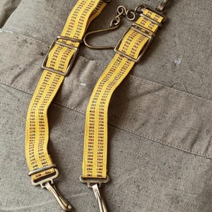 Op de afbeelding: Twee gele stoffen banden met een herhalend zwart cijferpatroon. Elke band is voorzien van verstelbare messingkleurige metalen schuifgespen en draaibare karabijnhaken aan beide uiteinden. Een decoratieve messingkleurige haak is aan een van de banden bevestigd.