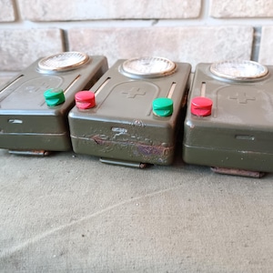 Peut inclure: Trois lampes de poche militaires vintage vert olive avec des boutons rouges et verts. Chaque lampe de poche a une lentille circulaire et un clip métallique en bas. Les lampes de poche sont disposées côte à côte sur une surface texturée.