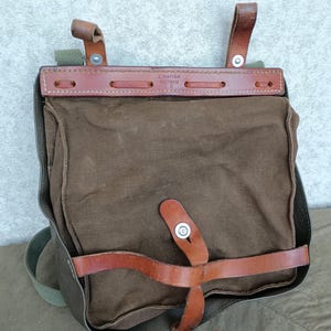 Puede incluir: Una bolsa de lona marrón con correas y detalles de cuero. La bolsa tiene forma rectangular con cierre de solapa y correa para el hombro. El texto "A MATTER REITNAU" es visible en la correa superior de cuero. La bolsa parece vintage.