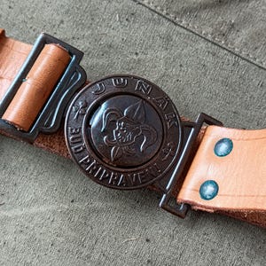 Könnte beinhalten: Ein hellbrauner Ledergürtel mit einer bronzefarbenen Schnalle. Die Schnalle hat ein kreisförmiges Design mit dem Wort "JUNAK" und einer Grafik eines Narren. Der Gürtel wird mit einem bronzefarbenen Metallverschluss befestigt.