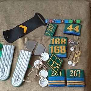 May include: A collection of military insignia including a black strap with a yellow chevron, silver shoulder boards, green and blue fabric patches with numbers 44, 168, 207, 287, a green diamond patch with crossed rifles, metal pins, and white circular tags.