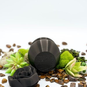 May include: A black ceramic rose and a black ceramic round object with a textured surface, surrounded by tiger's eye stones and green moss.