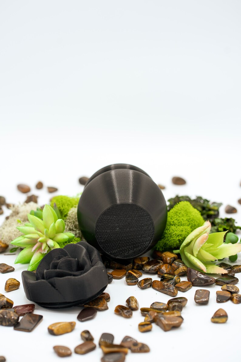 May include: A black ceramic rose and a black ceramic round object with a textured surface, surrounded by tiger's eye stones and green moss.
