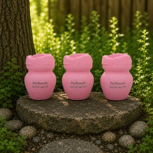 May include: Three pink urns with a flower-shaped lid are displayed on a stone. Each urn has the text "Kind Memorials" and dates. The urns are set against a backdrop of green foliage and a tree trunk.