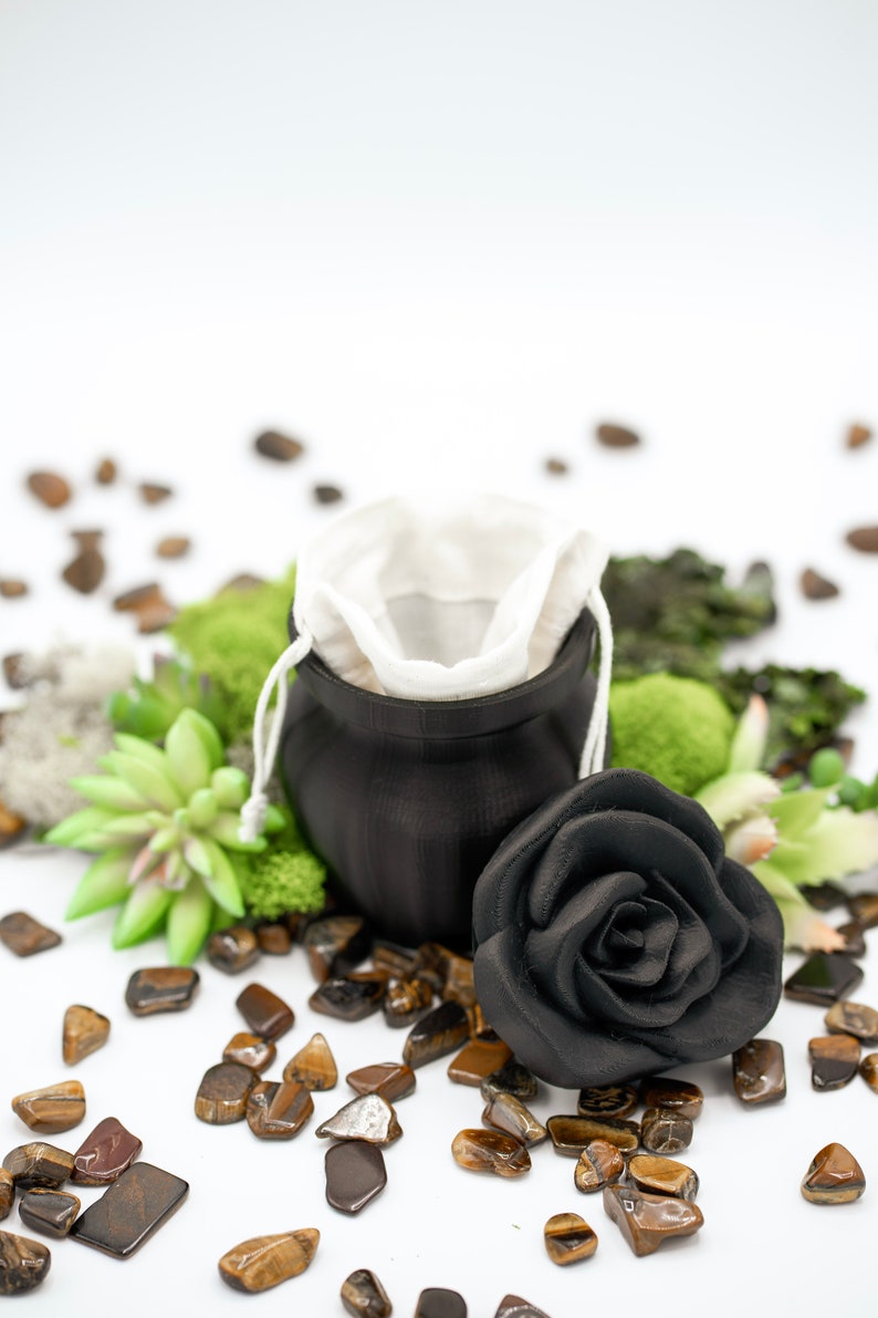 May include: A black ceramic jar with a white fabric pouch inside, surrounded by tiger's eye stones and a black rose.
