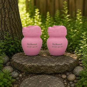 May include: Two pink urns with a flower-shaped lid, sitting on a stone. The urns have the text "Kind Memorials" and dates. The background features green foliage and a tree trunk.