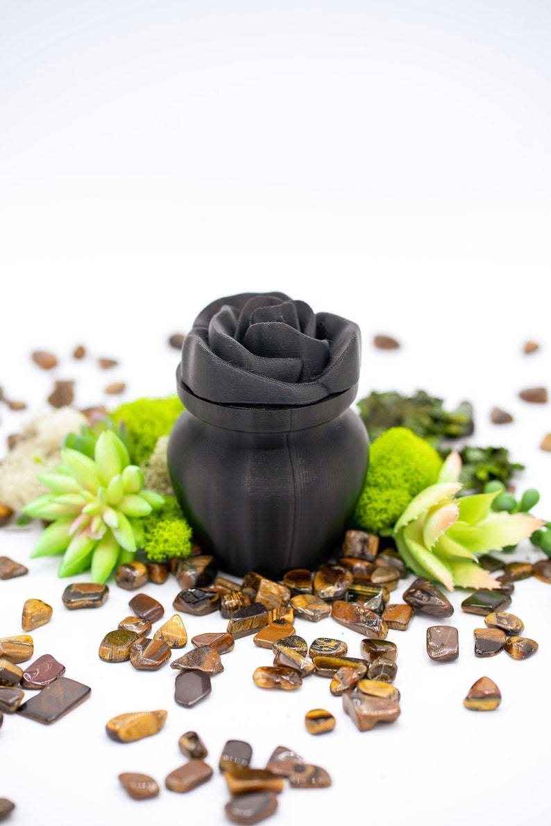 May include: A black rose-shaped cremation urn with a textured finish, surrounded by tiger's eye stones and green succulents.