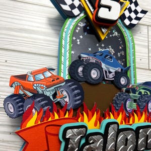 May include: A layered papercraft birthday decoration featuring monster trucks, flames, and checkered flags. The design includes a large number "5" and the name "Talon" in a bold, textured font. The color palette includes orange, blue, green, and black.
