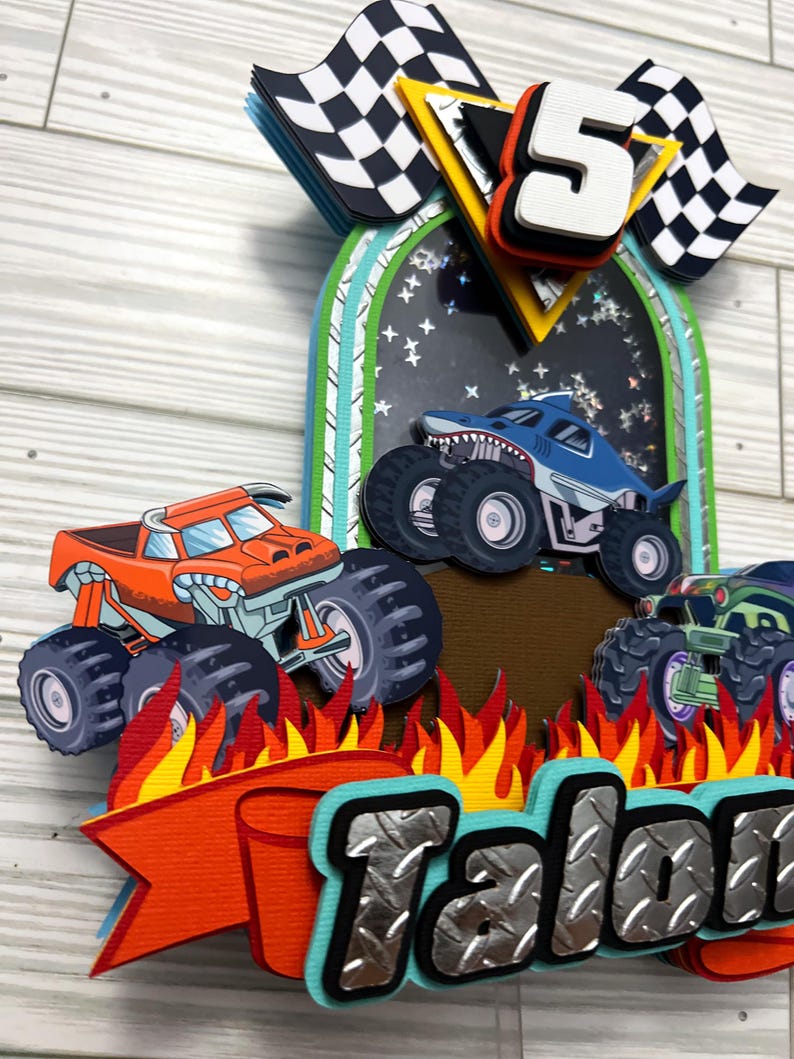 May include: A layered papercraft birthday decoration featuring monster trucks, flames, and checkered flags. The design includes a large number "5" and the name "Talon" in a bold, textured font. The color palette includes orange, blue, green, and black.