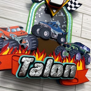 May include: A colorful monster truck-themed cake topper with three trucks, flames, and the name "Talon" in a diamond plate style. Includes a number 5 and checkered flags. The trucks are orange, blue, and green.