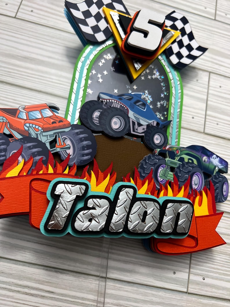 May include: A colorful monster truck-themed cake topper with three trucks, flames, and the name "Talon" in a diamond plate style. Includes a number 5 and checkered flags. The trucks are orange, blue, and green.