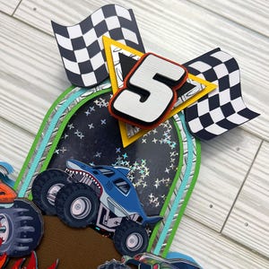 May include: A layered paper craft featuring a blue monster truck with a shark design, a large number 5, and checkered flags. The background includes a starry night design and a brown surface.