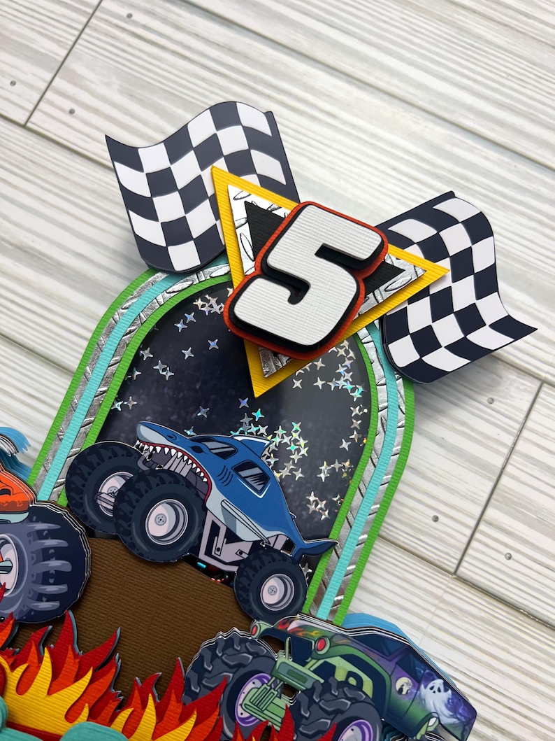 May include: A layered paper craft featuring a blue monster truck with a shark design, a large number 5, and checkered flags. The background includes a starry night design and a brown surface.