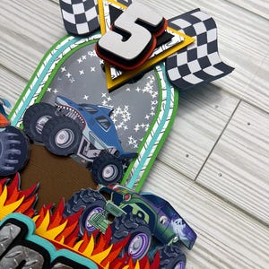May include: A layered paper craft featuring monster trucks, flames, and checkered flags. The number "5" is prominently displayed, suggesting a birthday theme. The design incorporates various colors, including blue, green, orange, and black, creating a dynamic and playful aesthetic.