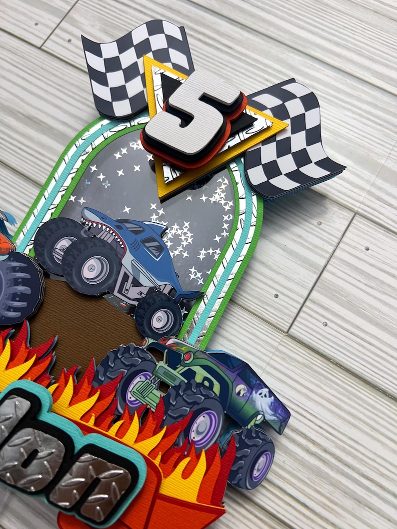 May include: A layered paper craft featuring monster trucks, flames, and checkered flags. The number "5" is prominently displayed, suggesting a birthday theme. The design incorporates various colors, including blue, green, orange, and black, creating a dynamic and playful aesthetic.