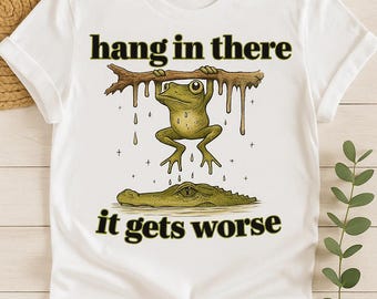 May include: White t-shirt with a humorous graphic. The design features a green frog hanging from a branch above a crocodile, with the text "hang in there it gets worse."