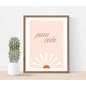 Könnte beinhalten: Ein gerahmter Druck mit den Worten "pura vida" in einer Schreibschrift, über einem stilisierten Sonnendesign. Der Hintergrund ist in einem weichen Pfirsichton gehalten. Links vom Rahmen befindet sich ein Kaktus in einem weißen Topf.