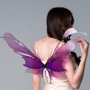 May include: Translucent purple and pink butterfly wings with black outlines. The wings are attached to a white metal frame. The wings are designed to be worn on the back. The wings are a decorative accessory.