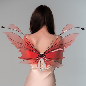May include: Red and pink translucent butterfly wings with black vein detailing. The wings are attached to a silver-coloured metal back piece. The wings are worn on the back.