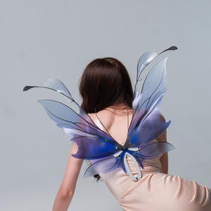 May include: A person wearing a peach-coloured dress with blue and purple butterfly wings. The wings have a translucent appearance with black outlines and are attached to the back of the dress. The person is leaning on their left arm.