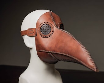 May include: A brown leather plague doctor mask with a long beak and eye holes. The mask has stitching around the edges and a small, round, perforated metal piece on the side.