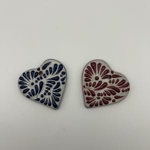 May include: Two heart-shaped ceramic ornaments. One is navy blue with white floral designs, the other is burgundy with white floral designs. Both have a hole at the top for hanging.