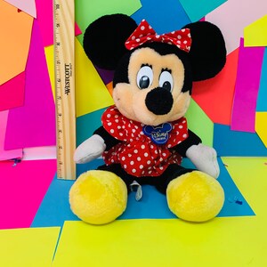 May include: A plush black and tan Minnie Mouse toy wearing a red dress with white polka dots and a red bow with white polka dots. The toy has a blue Disney Store tag attached to its dress. The toy is sitting on a colorful background.