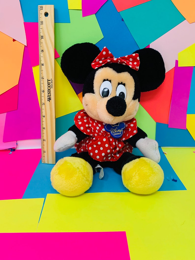 May include: A plush black and tan Minnie Mouse toy wearing a red dress with white polka dots and a red bow with white polka dots. The toy has a blue Disney Store tag attached to its dress. The toy is sitting on a colorful background.