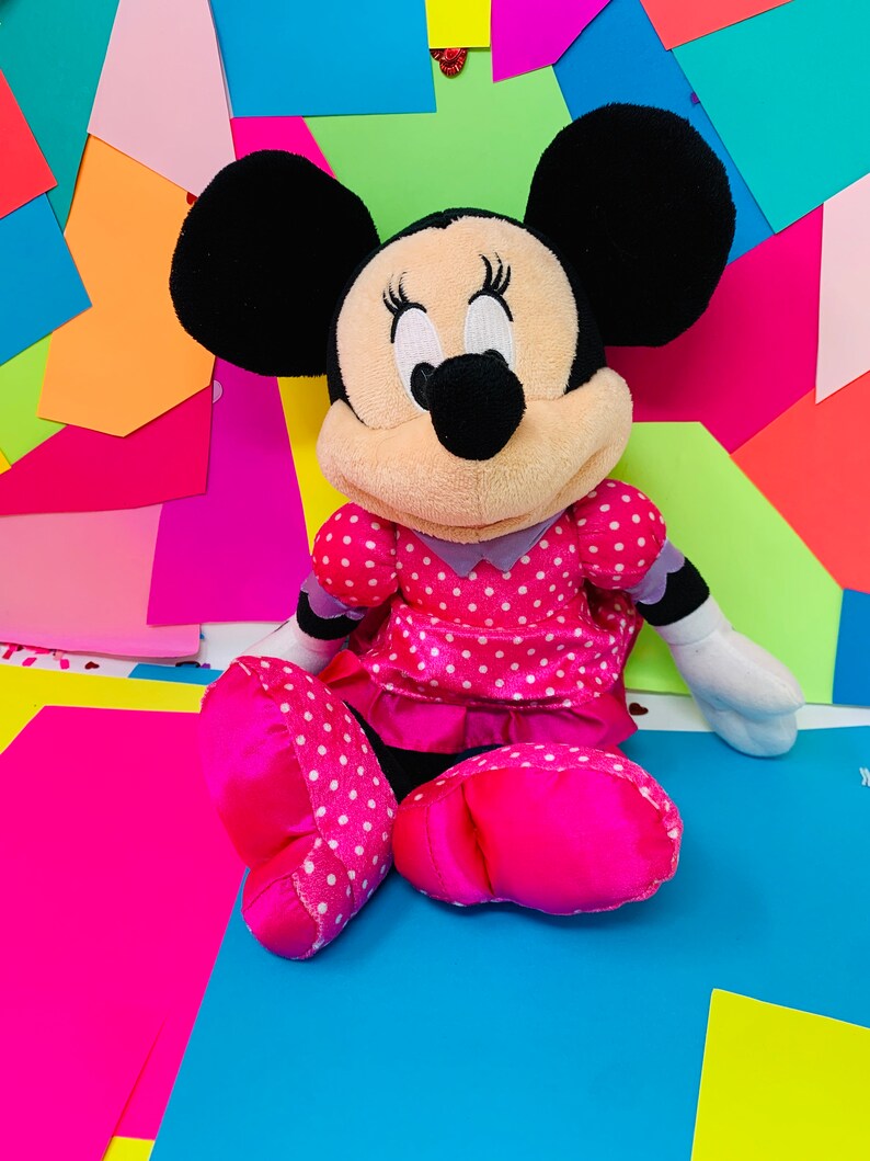 K&ouml;nnte beinhalten: Ein Pl&uuml;schtier von Minnie Mouse, die ein rosa Kleid mit wei&szlig;en Punkten tr&auml;gt. Sie hat schwarze Ohren und eine schwarze Nase. Das Spielzeug sitzt auf einem bunten Hintergrund.