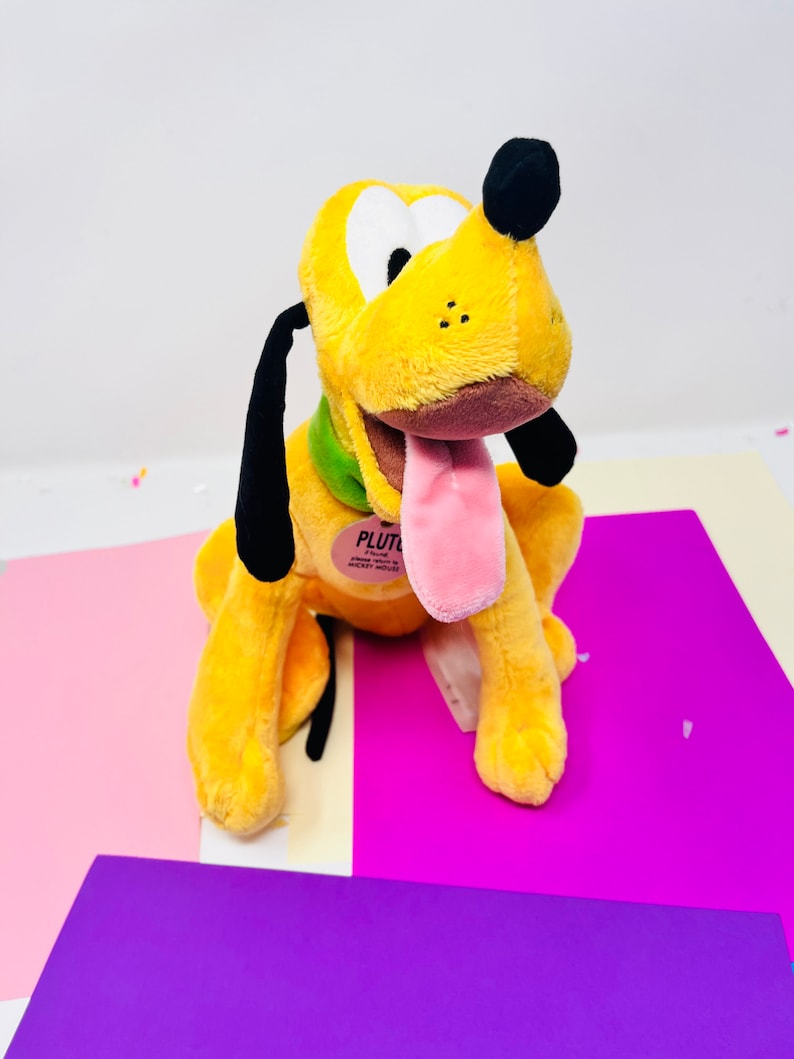 Pu&ograve; includere: Un cane di peluche giallo con orecchie nere e una lingua rosa che sporge. Il cane &egrave; seduto su una superficie viola e indossa un collare verde con la scritta "PLUTO" su di esso.