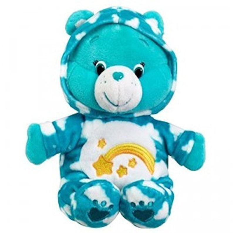 Pu&ograve; includere: Un peluche blu Care Bear che indossa un pigiama a motivi di nuvole bianche e blu con un arcobaleno e stelle. Il Care Bear ha un viso bianco con un naso rosa e occhi neri.