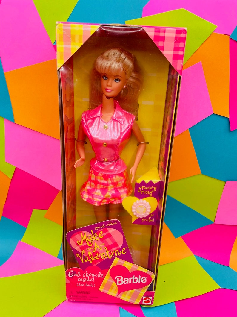 May include: A pink and yellow Barbie doll box with a pink and white plaid pattern. The box features the text "Make A Valentine" and "Groovy Thing" with a pink heart. The doll is wearing a pink shirt, a plaid skirt, and a gold bracelet.