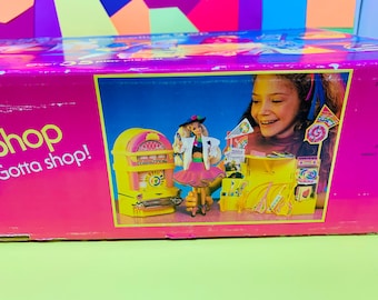 May include: A pink and yellow box with a Barbie doll and a jukebox. The box says "Shop! Gotta shop!" and features a girl with a yellow headband.