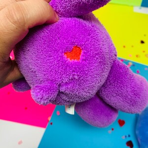 Pu&ograve; includere: Peluche viola con un cuore rosso ricamato sulla schiena. Il giocattolo &egrave; tenuto da una mano.