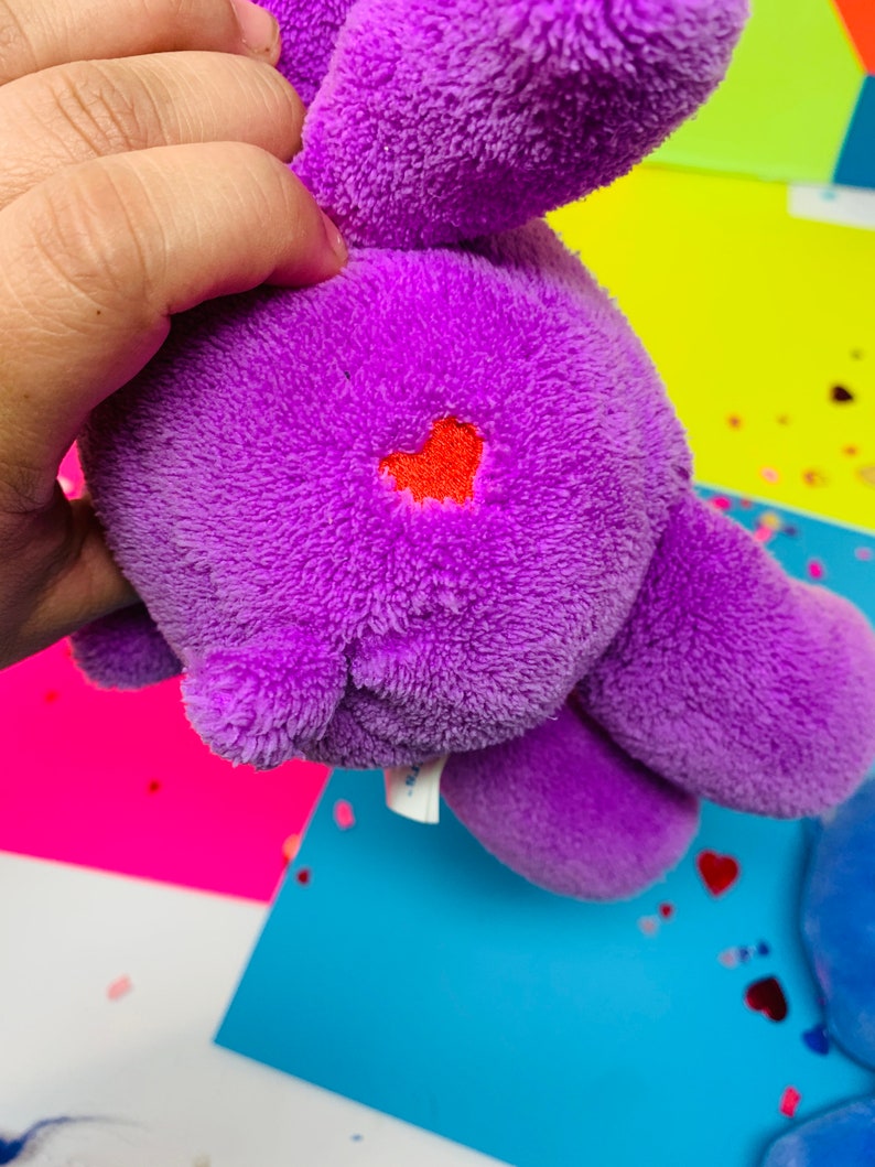 Pu&ograve; includere: Peluche viola con un cuore rosso ricamato sulla schiena. Il giocattolo &egrave; tenuto da una mano.