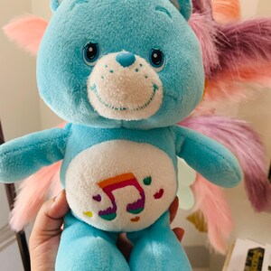 Pu&ograve; includere: Un orsacchiotto di peluche blu Care Bear con una nota musicale arcobaleno sul pancino. L'orso ha un viso bianco con occhi neri e un naso rosa.