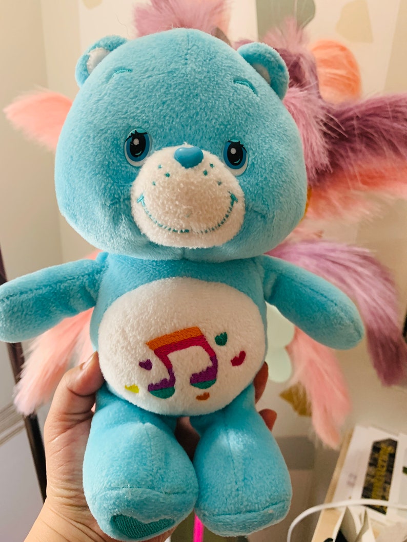 Pu&ograve; includere: Un orsacchiotto di peluche blu Care Bear con una nota musicale arcobaleno sul pancino. L'orso ha un viso bianco con occhi neri e un naso rosa.