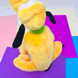 Pu&ograve; includere: Un cane di peluche giallo con orecchie nere e un collare verde. Il cane &egrave; seduto su una superficie viola.