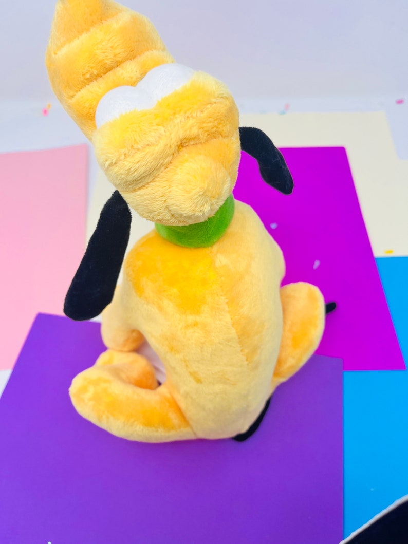 Pu&ograve; includere: Un cane di peluche giallo con orecchie nere e un collare verde. Il cane &egrave; seduto su una superficie viola.