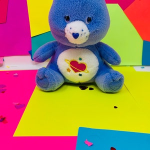 Pu&ograve; includere: Un orsacchiotto di peluche blu con la pancia bianca e un cuore rosa con un pianeta intorno. L'orso &egrave; seduto su una superficie gialla con carta rosa e blu dietro.