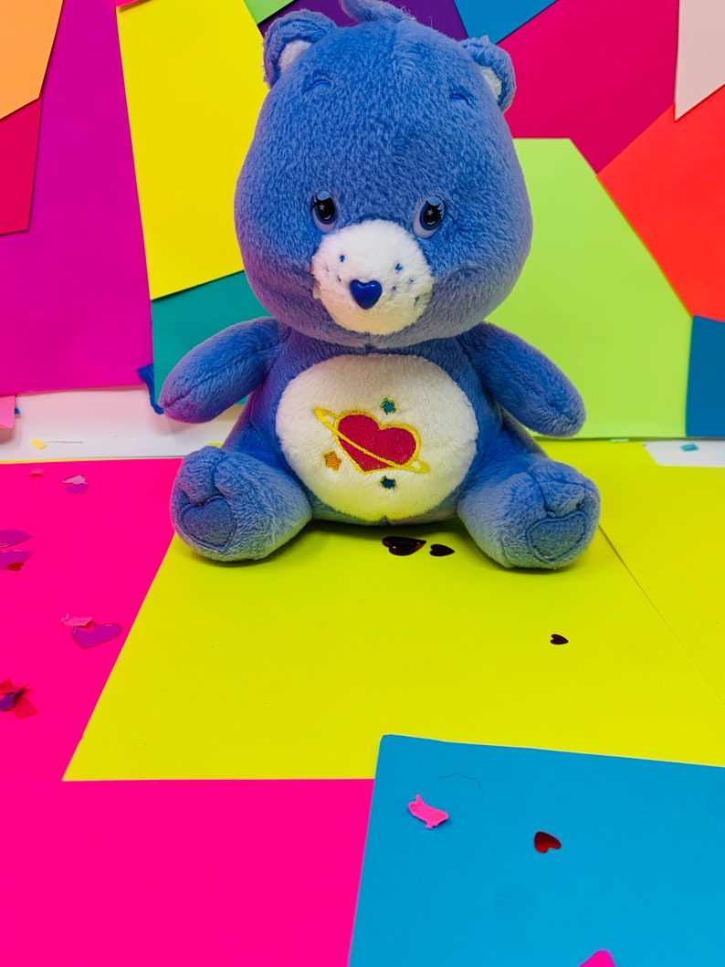 Pu&ograve; includere: Un orsacchiotto di peluche blu con la pancia bianca e un cuore rosa con un pianeta intorno. L'orso &egrave; seduto su una superficie gialla con carta rosa e blu dietro.