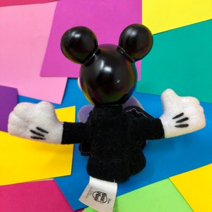Op de afbeelding: Zwart pluche Mickey Mouse speelgoed met witte handschoenen en een zwart en paars outfit. Het speelgoed zit op een kleurrijke achtergrond met de tekst "Disney" en "Kohl's Kids" op het label.