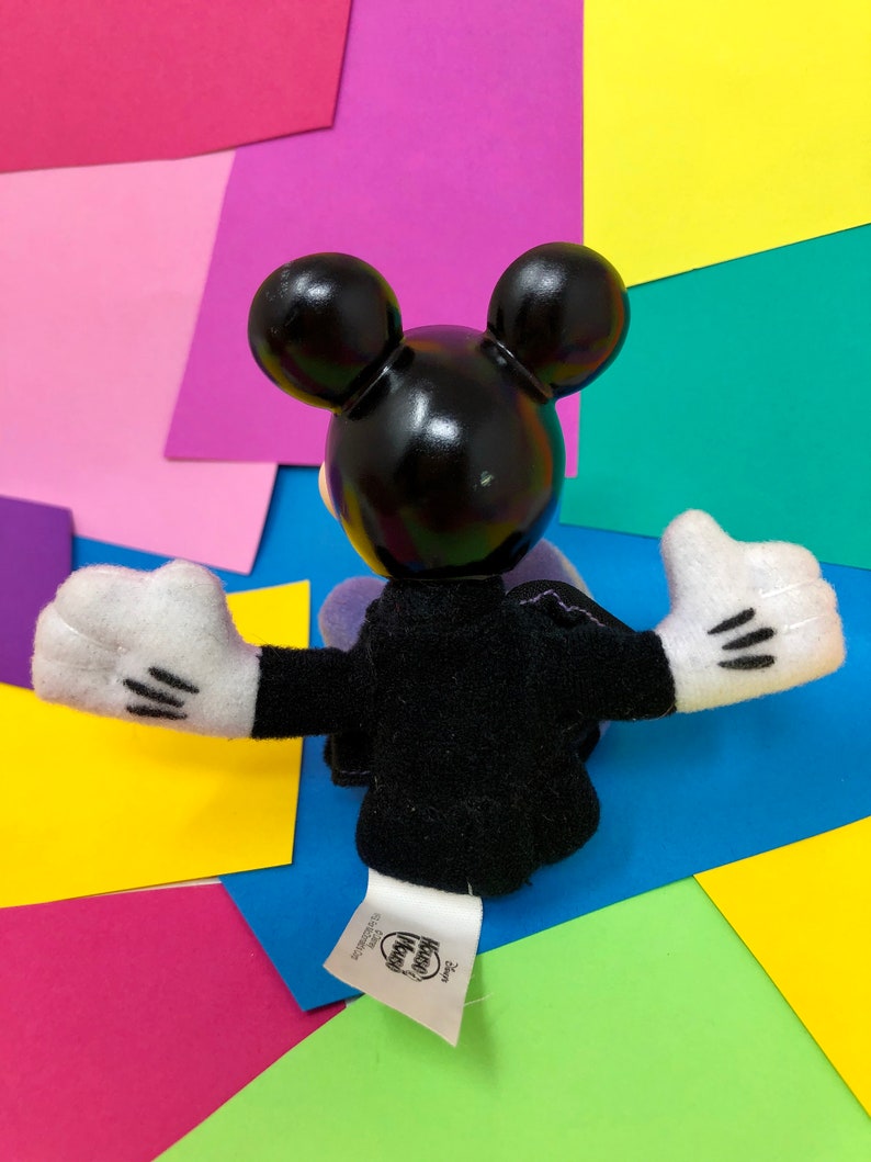 Op de afbeelding: Zwart pluche Mickey Mouse speelgoed met witte handschoenen en een zwart en paars outfit. Het speelgoed zit op een kleurrijke achtergrond met de tekst "Disney" en "Kohl's Kids" op het label.
