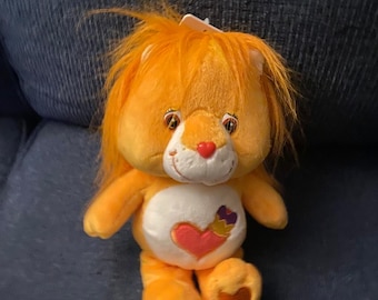 May include: Orange Care Bear plush toy with a heart and crown on its chest. The bear has a long mane of orange fur and a white face with red cheeks.