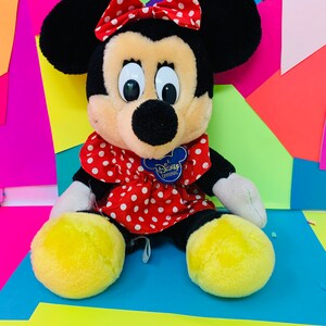 May include: A plush Minnie Mouse toy wearing a red dress with white polka dots and a red bow with white polka dots. The toy has a yellow Disney Original tag on its chest.