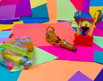 May include: A set of three plastic toys from a Happy Meal. The toys are a brown dog with a blue spot on its head, a green and yellow toy with a face, and a red and yellow toy of a man lifting weights. The toys are on a colourful background of pink, purple, yellow, and green paper.