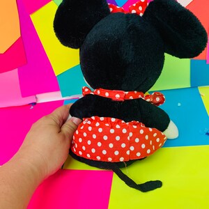 May include: A black and red plush toy mouse wearing a red polka dot dress and a red bow. The toy is sitting on a colorful background.